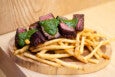 Sliced steak with a green chimichurri-like sauce, over skinny fries, on a light wooden platter.