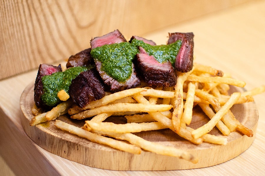 Sliced steak with a green chimichurri-like sauce, over skinny fries, on a light wooden platter.