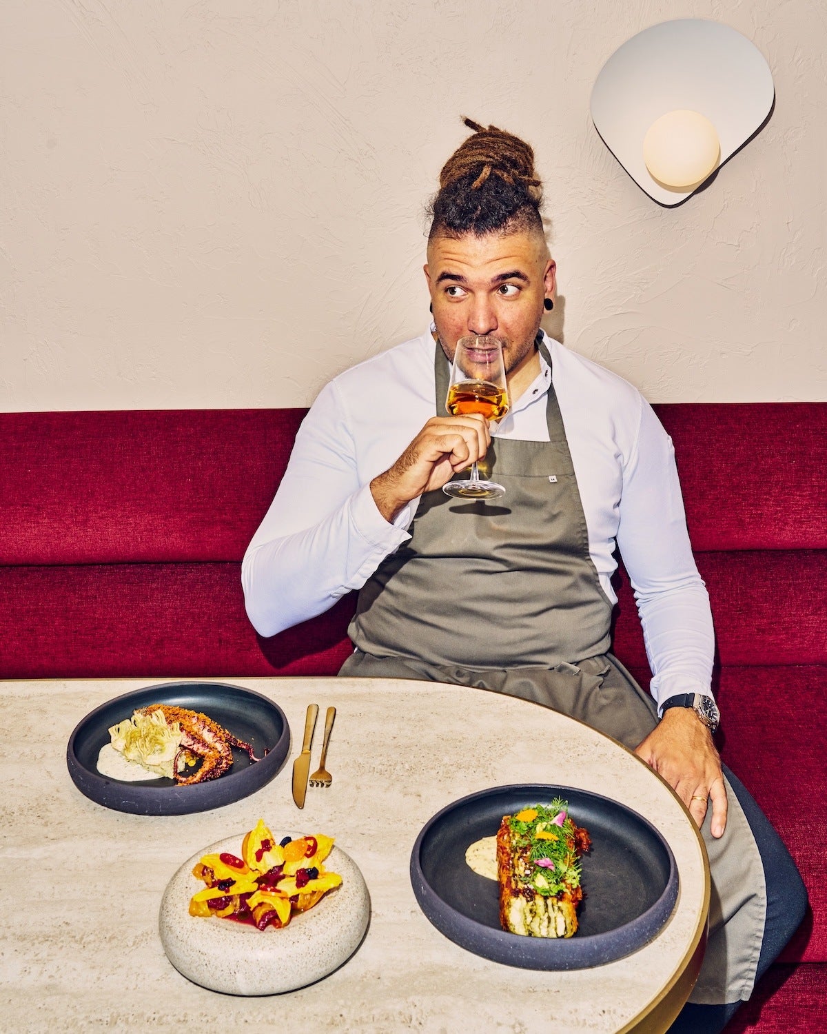 A man in a chef apron sits in a red booth, sipping a glass of wine, with modern Greek dishes on the table in front of him.