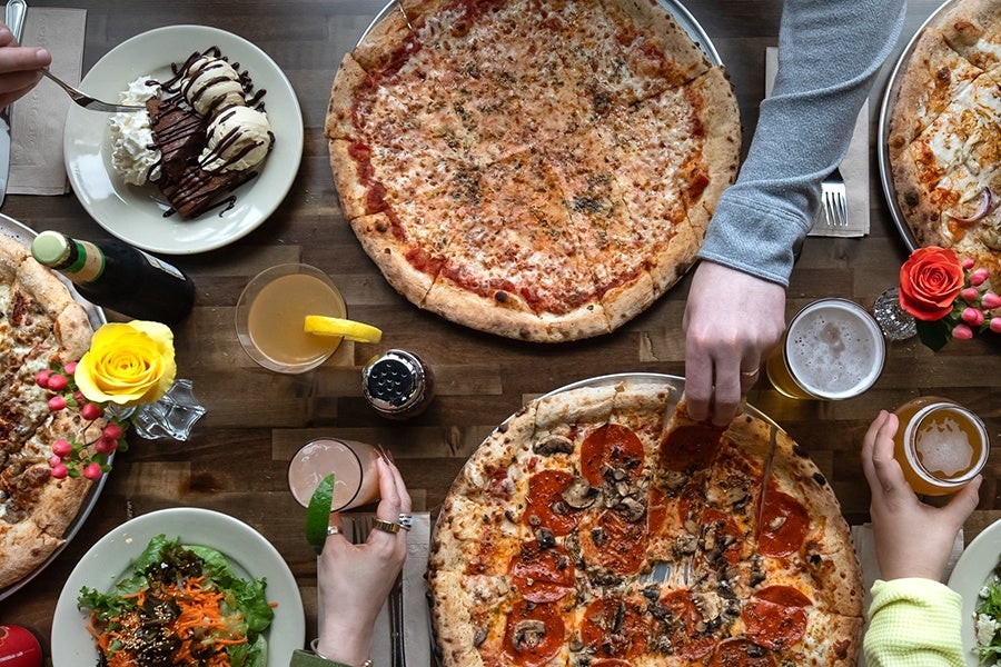 Overhead view of a table with pizzas, salad, and a brownie sundae, with hands reaching in to grab food and beer.
