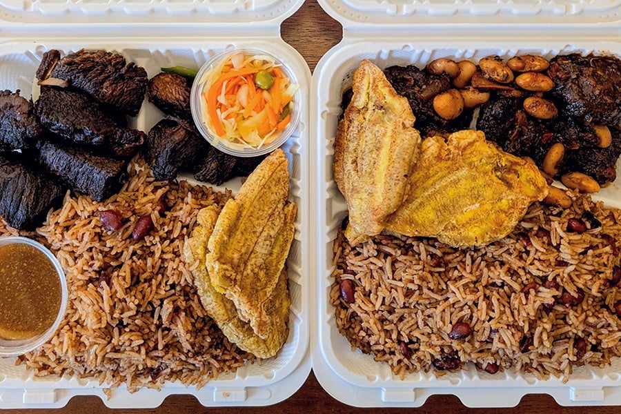 Overhead view of two takeout containers of Haitian food, including fried beef and oxtail with rice and plantains.