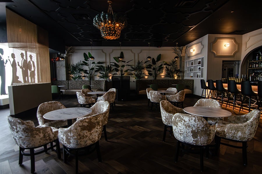 Interior of a bar with champagne-colored, crushed-velvet chairs, greenery, and a coconut-shell chandelier. Life-sized decals of 1920s-dressed silhouettes are visible on the window.
