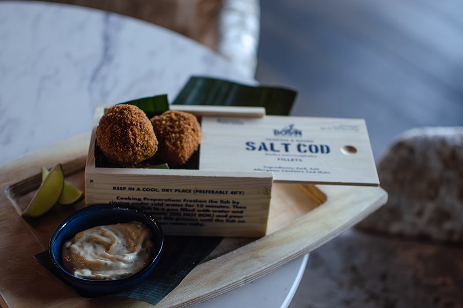 Crispy fried balls are displayed in a wooden salt cod box on a white marble table.
