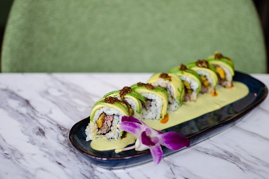 An avocado-wrapped sushi roll is stuffed with steak and plantain, displayed on a white marble table.