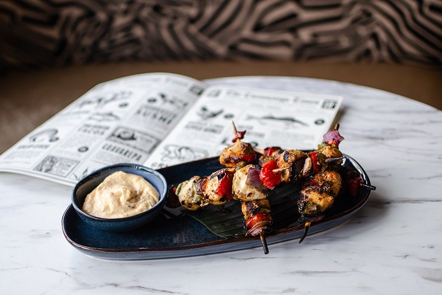 Grilled chicken and peppers on skewers are displayed on a white marble table with a thick off-white dipping sauce.