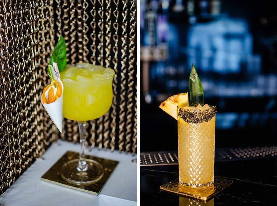 Two tropical cocktails, one bright yellow with a basil leaf and one pale orange with a wedge of pineapple and a black salted rim.