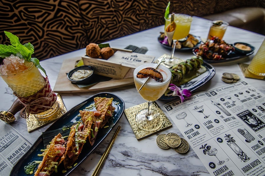 Spread of Latin-Asian fusion dishes, tropical cocktails, and novelty golden coins on a white marble table.