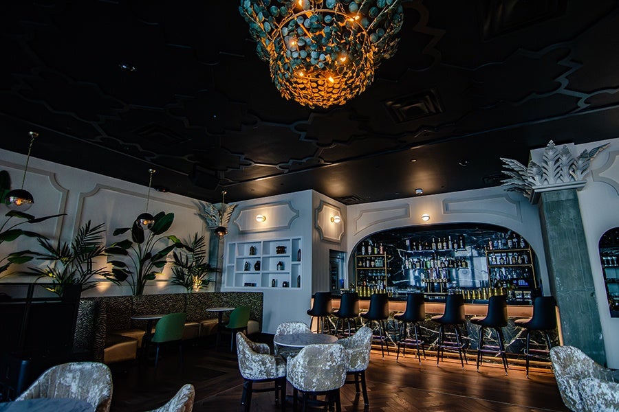 Interior of a bar with champagne-colored, crushed-velvet chairs, greenery, and a coconut-shell chandelier. Decorative pillars are topped with palm frond sculptures.