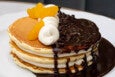 A stack of pancakes is topped with chocolate sauce, chocolate chips, a swirl of ricotta, and orange slices