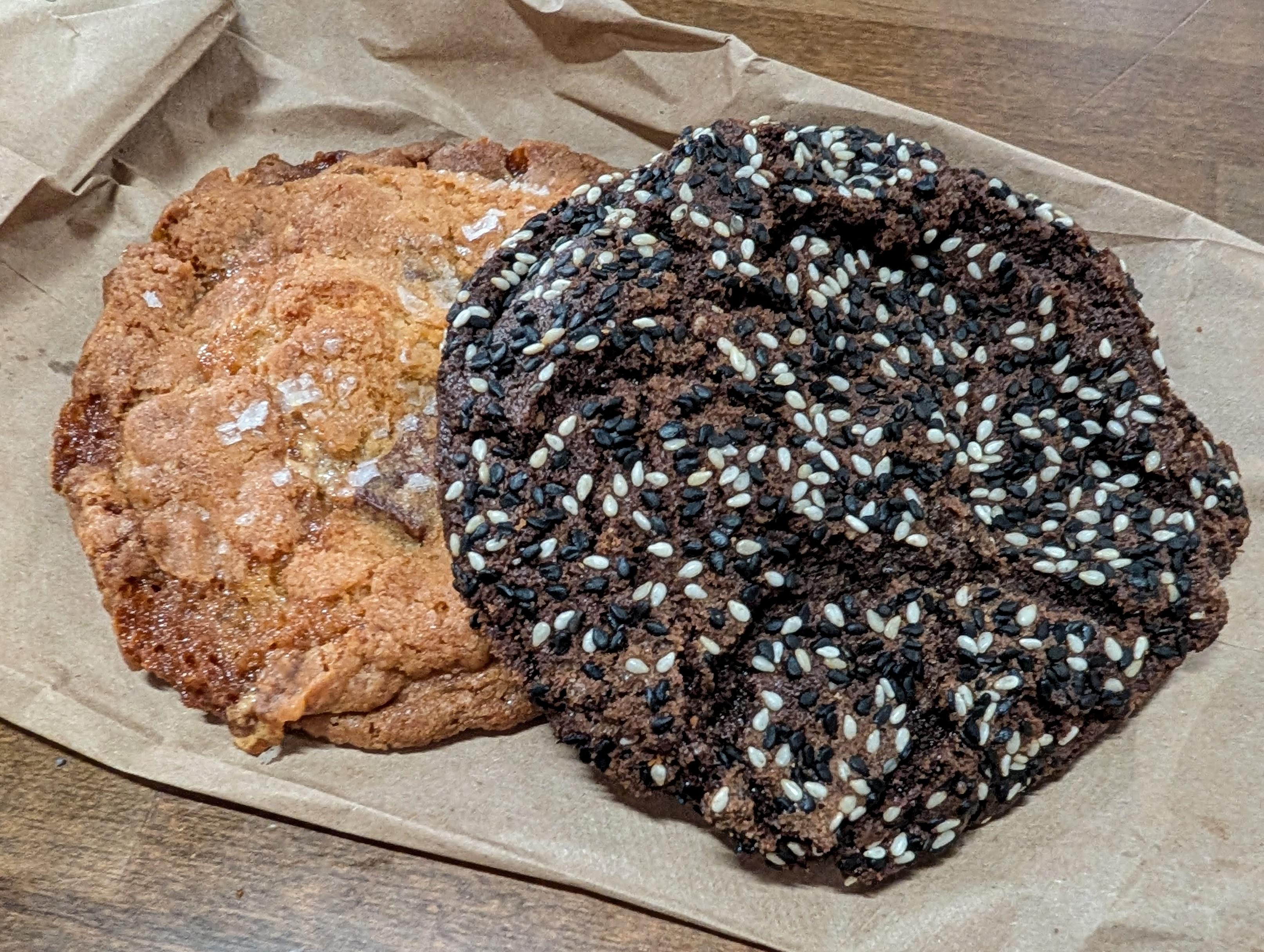 Two big cookies sit on a paper bag. One is chocolate and covered with sesame seeds, while the other has bits of marshmallow and other fillings.