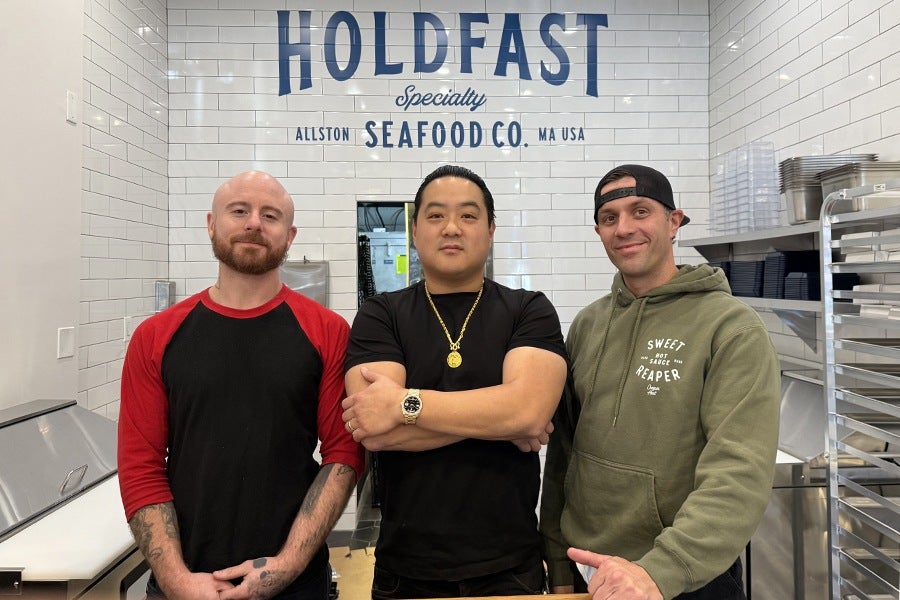 Three men stand in front of a white subway-tile wall with lettering that says Holdfast Specialty Seafood Co. Allston MA USA.