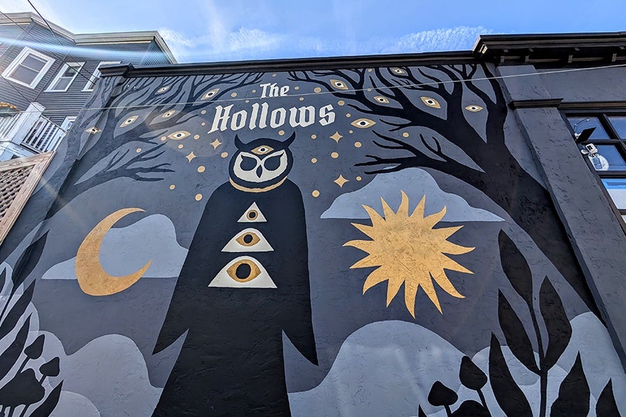 A mural on the side of the building says The Hollows and features an owl figure with a moon, sun, and trees.