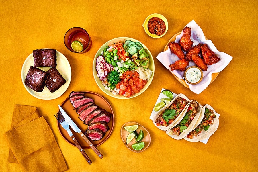 Overhead view of steak, brownies, salad, wings, tacos, and accoutrements on a yellow-orange background.