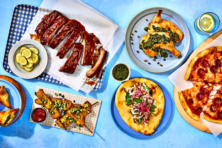 Overhead view of wings, pizza, chicken with a green sauce, a pita, and more on a blue background.