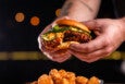 Two hands hold a saucy, crispy fried chicken sandwich, topped with pickles, over a pile of tater tots.