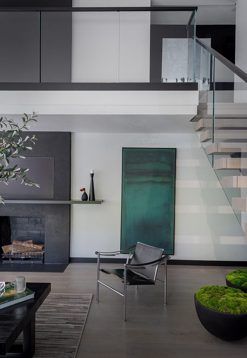 Modern living room with a black leather and chrome chair, a large green abstract painting on the wall, a black fireplace with logs, a floating shelf holding two decorative vases, a glass and wood staircase with a glass railing, and two large black planters filled with green moss. The space features a neutral color palette with gray, black, and white tones.