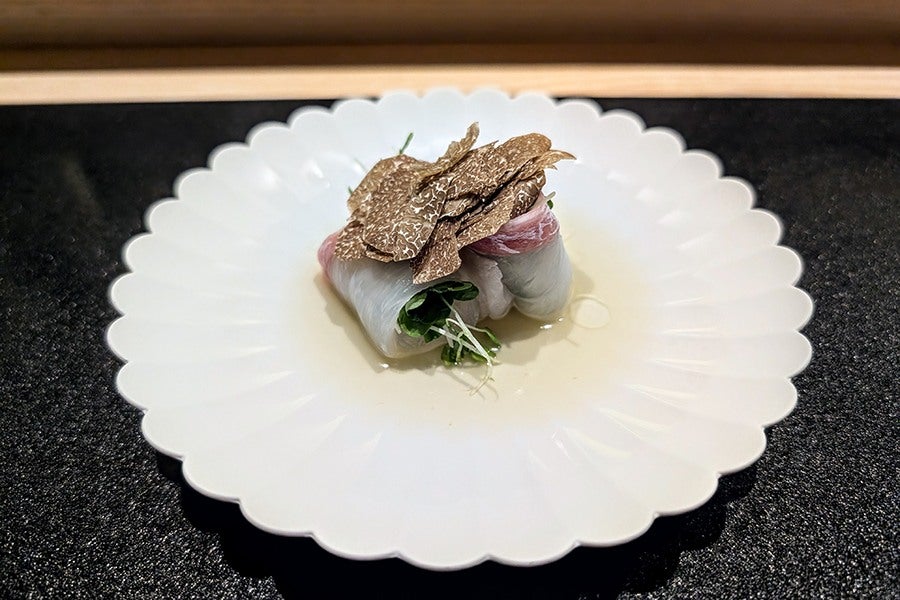 Several pieces of a raw white fish are rolled up with microgreens and a light dressing, topped with ample black truffle slices.