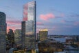 Photographed at sunset, a skyscraper with reflective windows rises over a cityscape, with water in the distance.