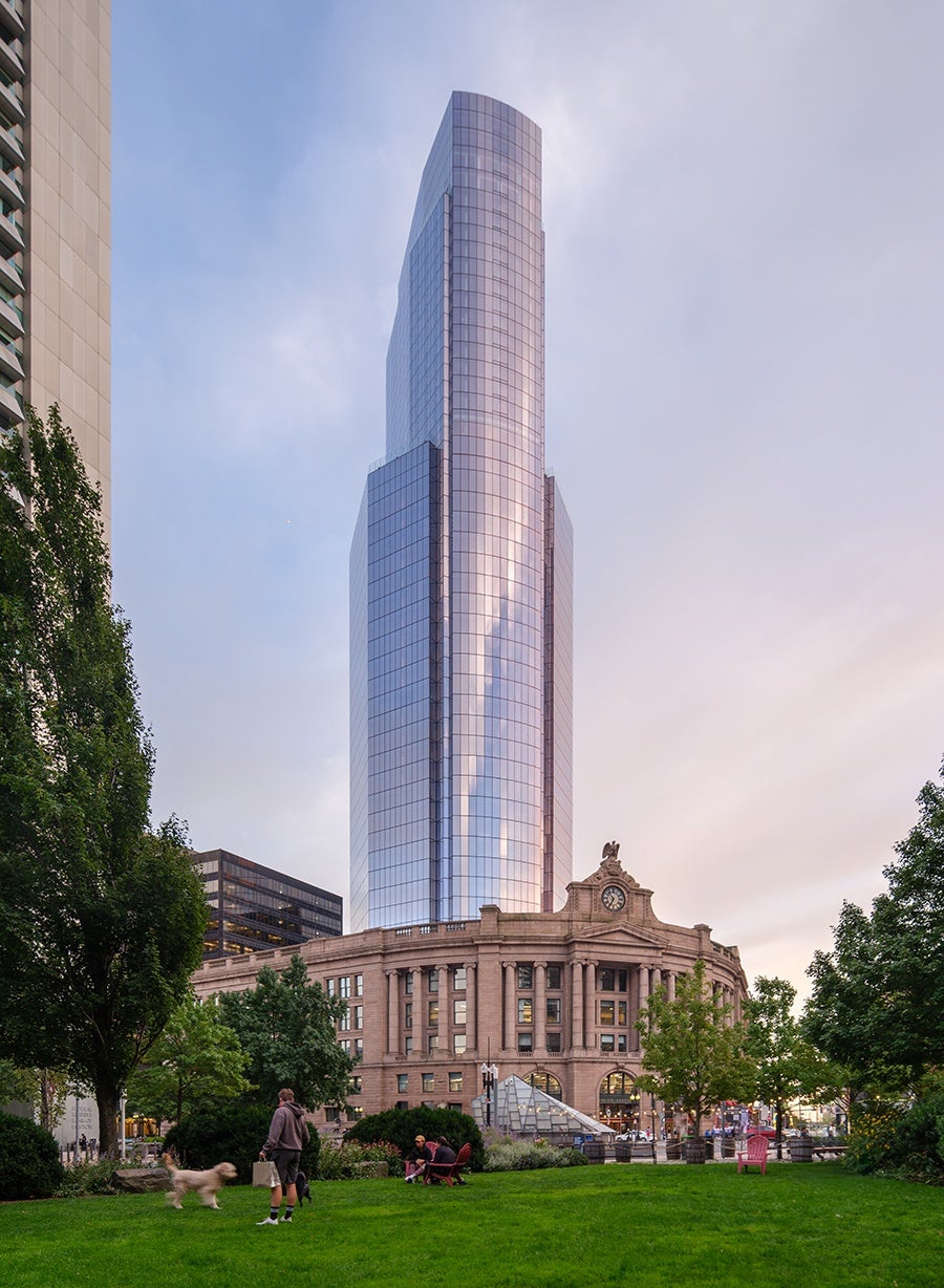 A shining skyscraper rises up over Boston's historic granite South Station, photographed from the grassy Dewey Square across the street.