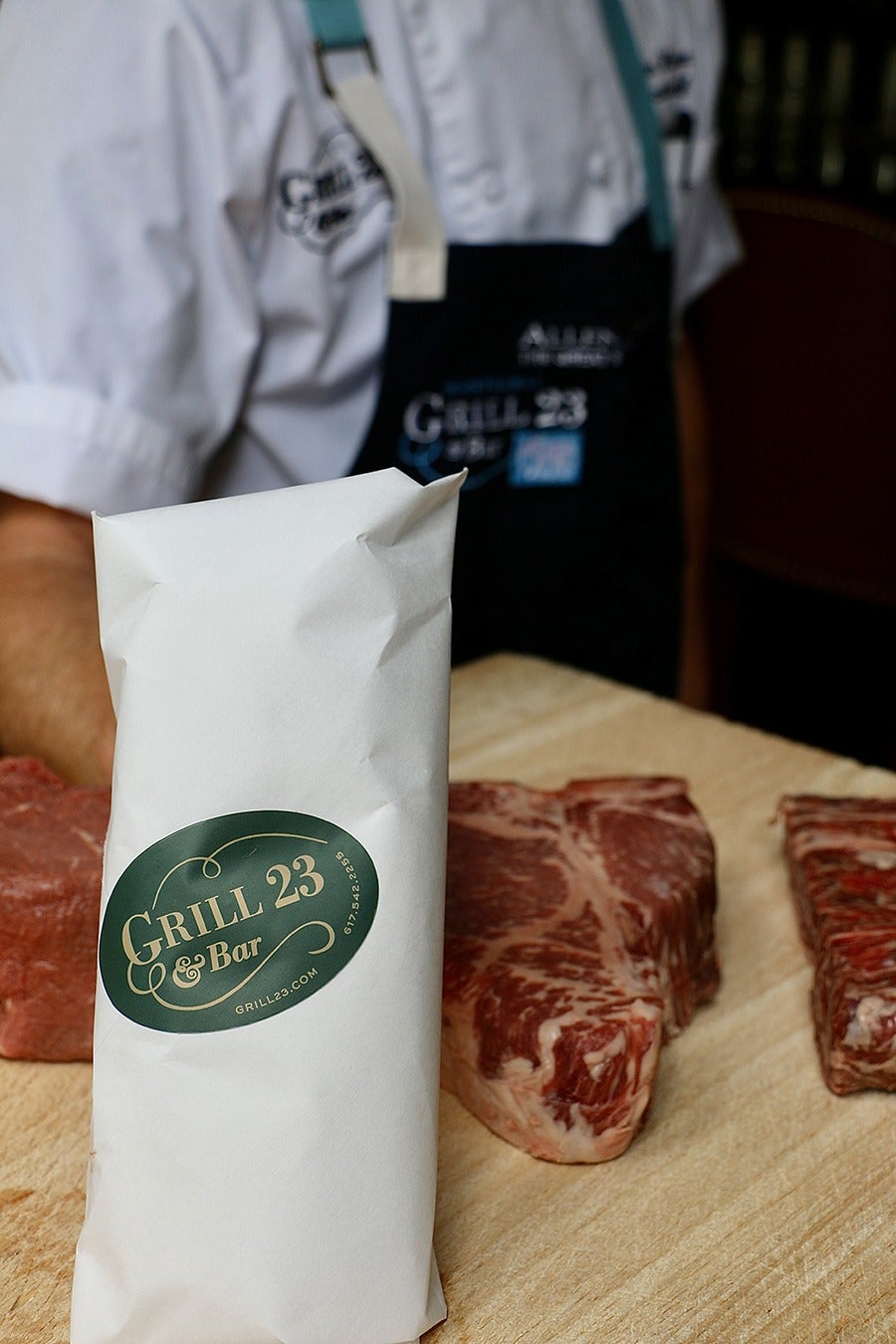 Raw steaks on a cutting board next to a white paper-wrapped object with a Grill 23 sticker on it.
