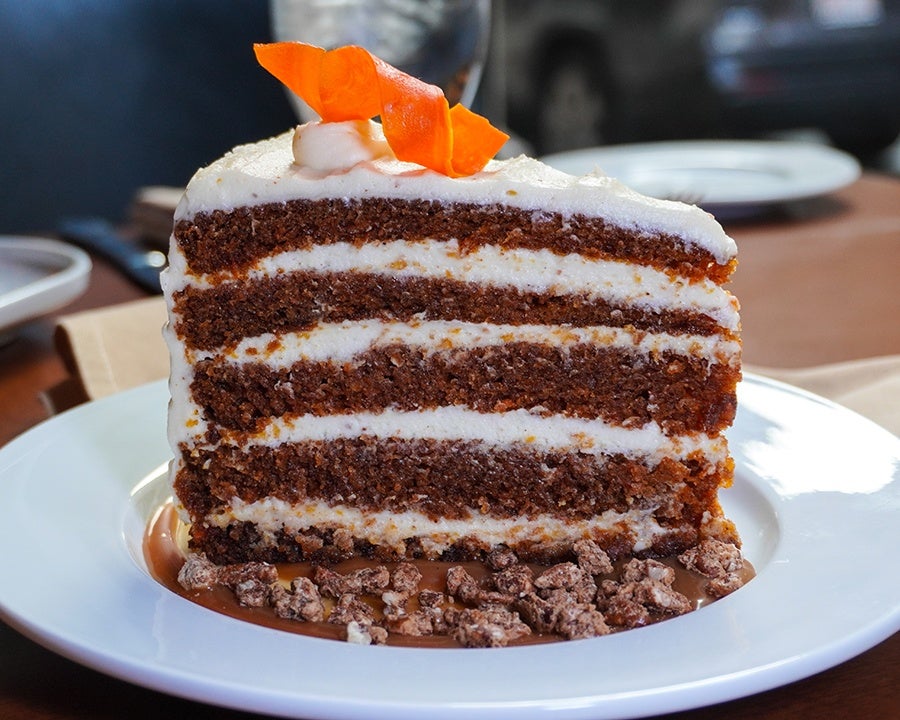 A big slice of carrot cake sits in a pool of caramel with candied nuts and is garnished with a spiral of carrot.