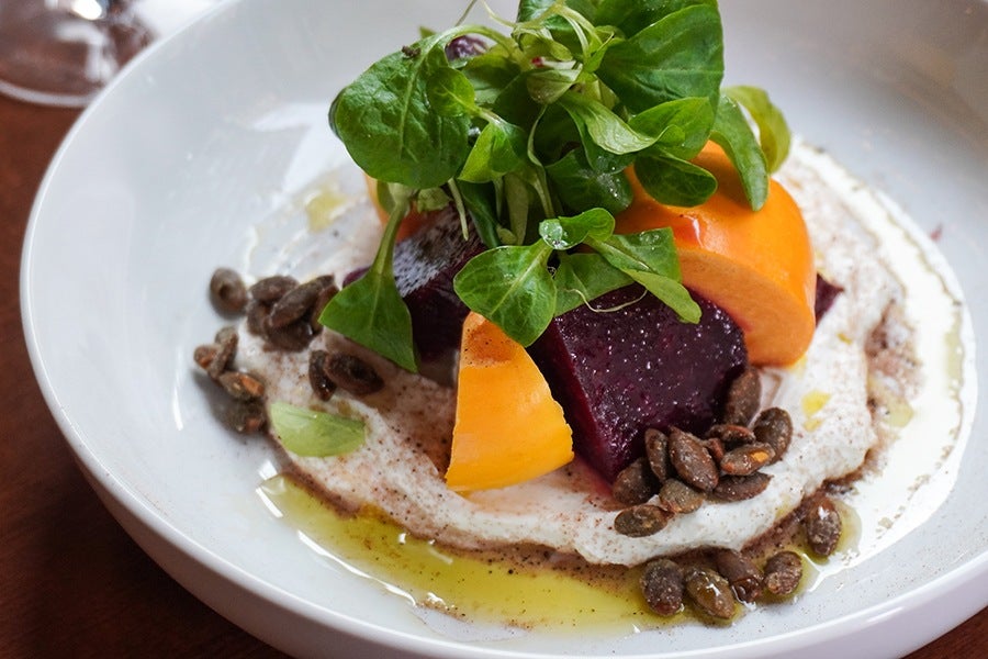 Big slices of purple beets and orange persimmon sit on a bed of white, creamy labneh, garnished with greens and pumpkin seeds.