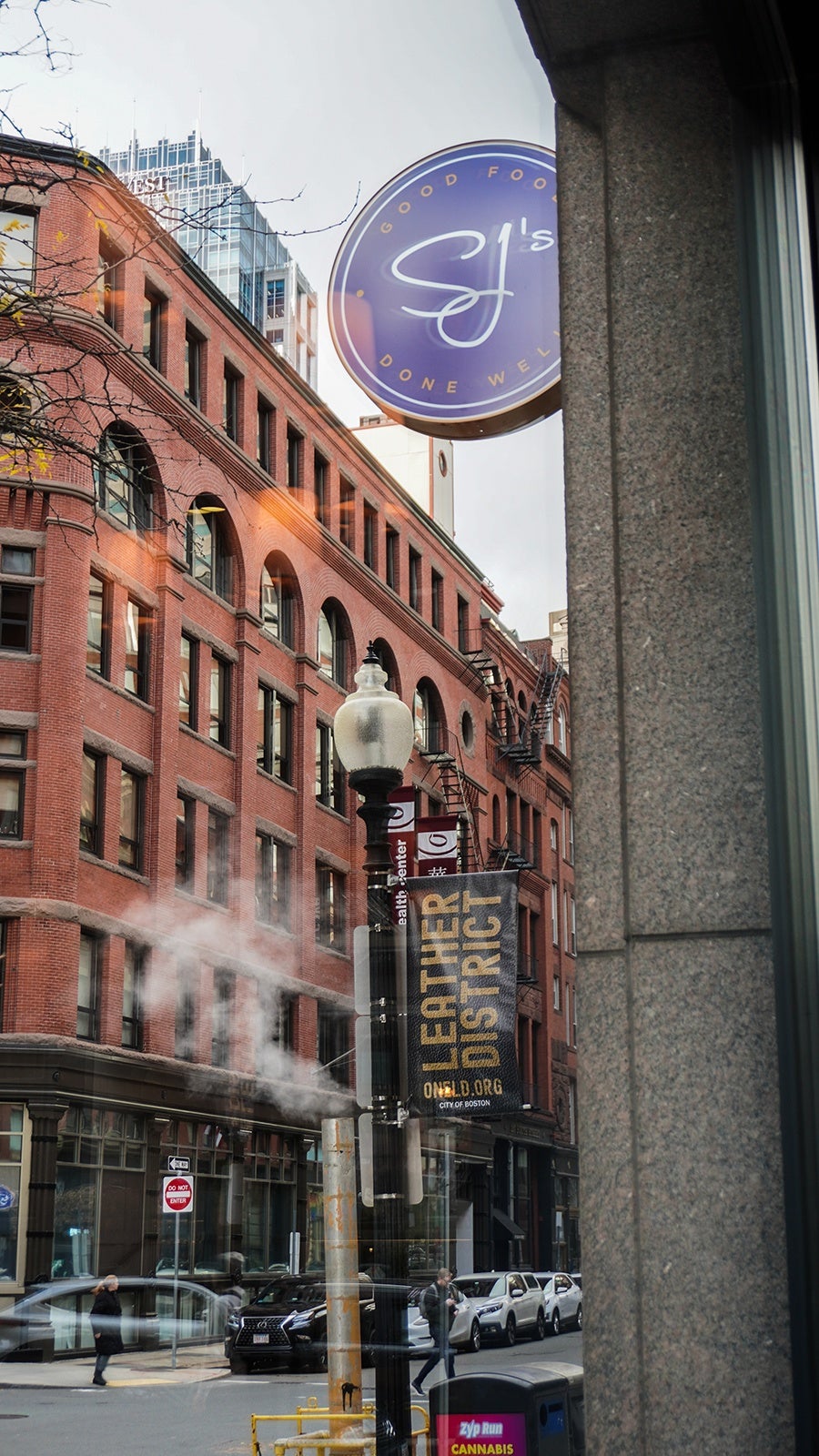 Photographed through a restaurant window from the inside, the restaurant's sign is visible attached to a building on a city street. The sign says SJ's - good food done well.