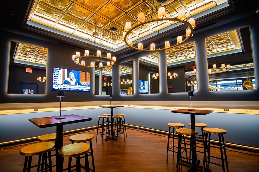 A corner of a restaurant, empty of patrons, features high-top tables, counter space, a television, and dark blue and gold decor.