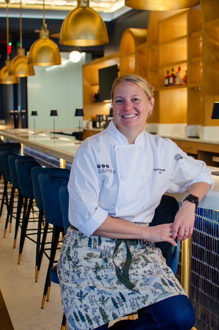 A chef in a white coat and pine tree-covered apron, her blond hair pulled back in a bun, sits at a bar in an empty restaurant, smiling at the camera.