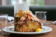 A lobster roll on a toasted bun is dripping with butter and served with chips and a lemon wedge.
