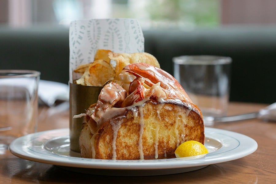 A lobster roll on a toasted bun is dripping with butter and served with chips and a lemon wedge.
