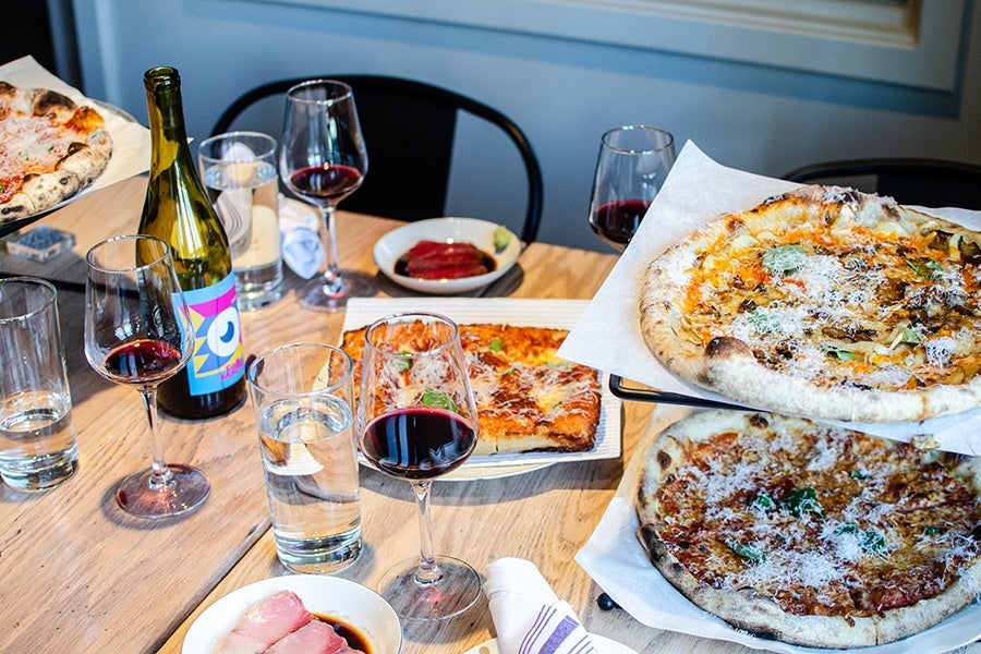 Wood-fired pizzas—some round, one square—sit on a light wooden table with a couple plates of simple sashimi and glasses of red wine.