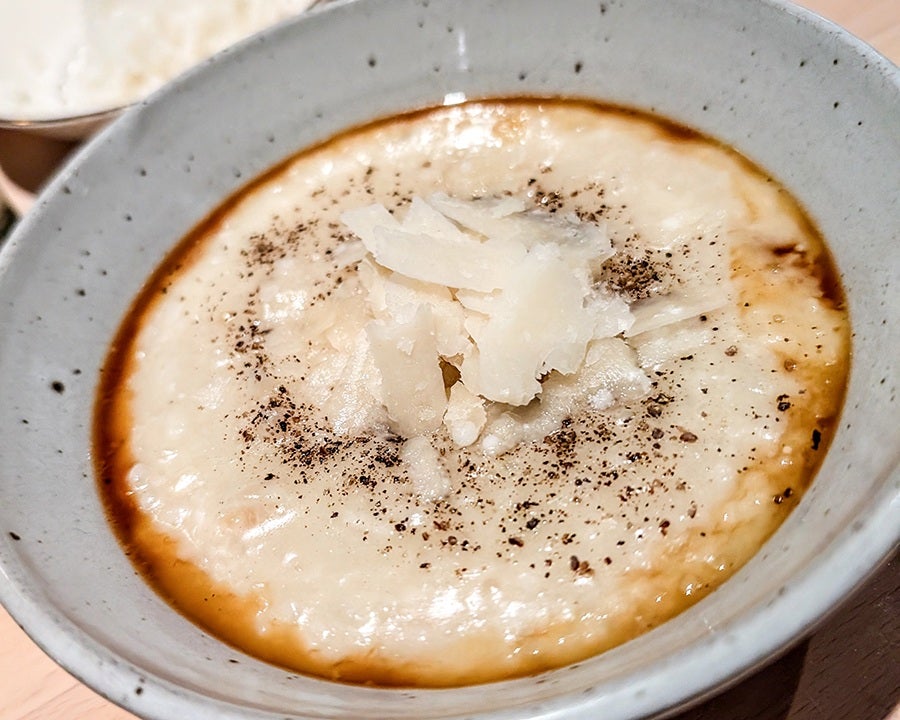 Closeup on a bowl of risotto with a ring of a brown broth around the edges. It's topped with chunks of parmesan and generous black pepper.