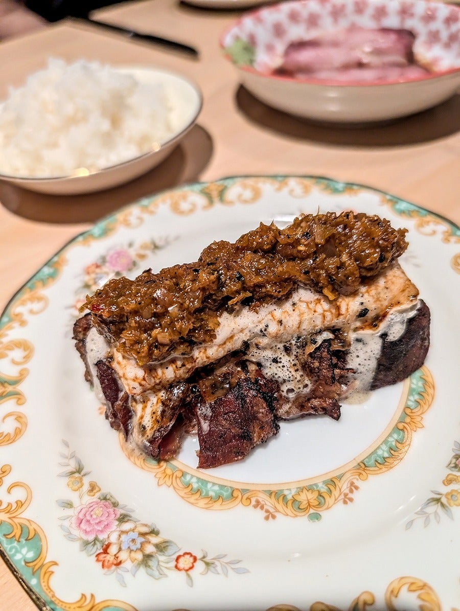 A big hunk of pork belly sits on a vintage floral plate, topped with a white sauce and an orange-brown chutney.