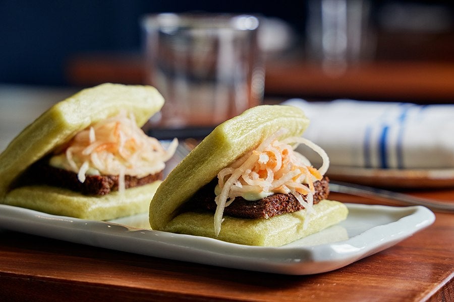 Two soft bao buns, tinted light green, are stuffed with a fried patty and pickled vegetables.