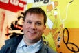 A man with short brown hair and blue eyes is smiling at the camera. He is wearing a light blue collared shirt under a dark jacket. Behind him is a colorful wall with a yellow and orange cartoon illustration of a person riding a bicycle.