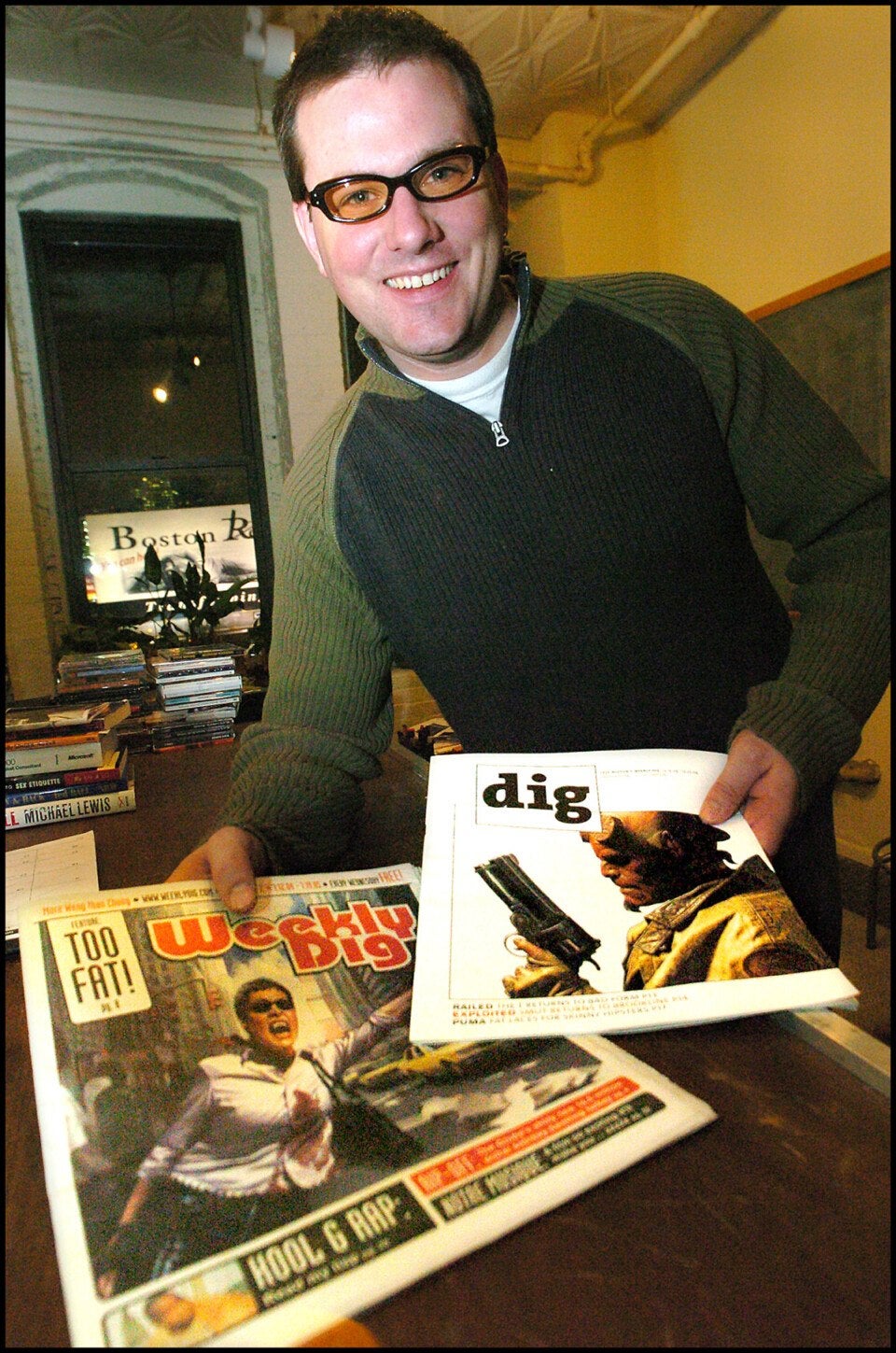A man wearing glasses and a green and black sweater is smiling and holding two magazines. One magazine is titled "Weekly Dig" with a cover image of a person running and the headline "TOO FAT!" The other magazine is titled "dig" with a cover image of a person holding a gun. The background shows a window, a stack of books, and a yellow wall.