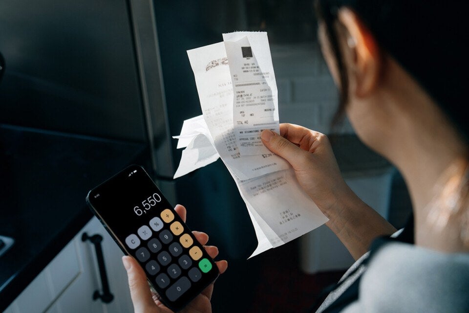 A person holding several receipts in one hand and a smartphone with a calculator app open displaying the number 6,550 in the other hand. The person is viewed from behind, focusing on the hands and the items held.