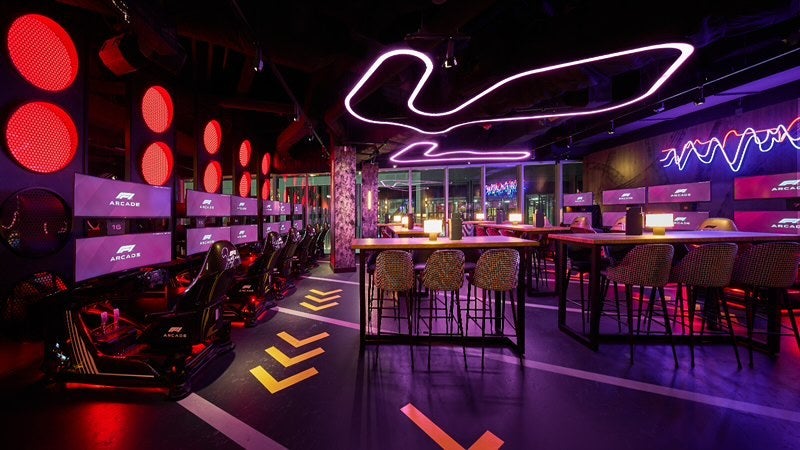A dark room lit by neon of barstools, racing videogames, and tables.