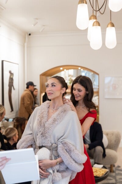 Two women are in the foreground, with one wearing a light gray fur-trimmed coat and the other in a red dress fastening a necklace around the first woman's neck. The setting appears to be a cozy, well-lit room with modern decor, including a large mirror with wooden framing and a chandelier with white and brass light fixtures. Several people are in the background, engaged in conversation.