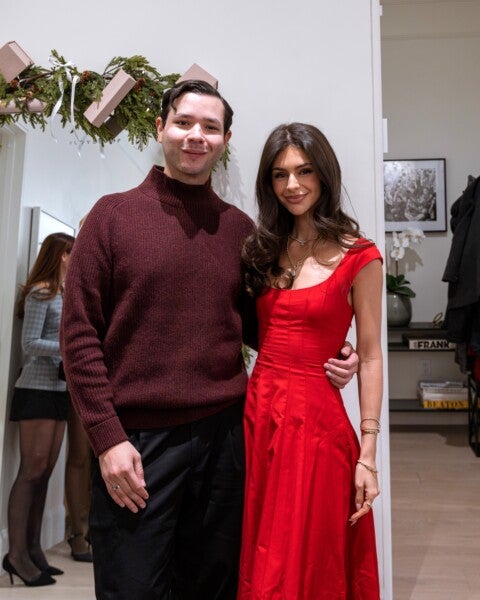 A man and a woman standing close together indoors. The man is wearing a maroon ribbed sweater and black pants, while the woman is dressed in a red fitted dress with cap sleeves. The woman has long, wavy dark hair and is accessorized with gold jewelry. Behind them, a mirror reflects another woman wearing a gray plaid blazer, black skirt, and black heels. The background includes a decorated garland with pinecones and pink boxes, a framed black-and-white photo, a white orchid plant, and some books on a shelf.