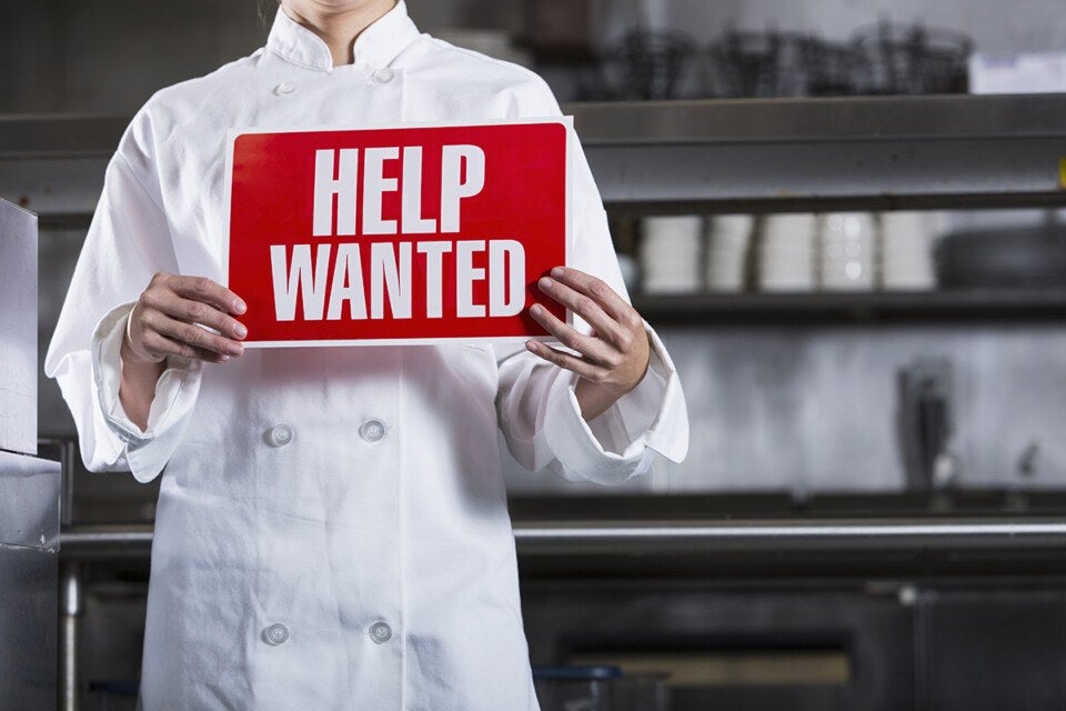 Person wearing a white chef's coat holding a red sign with white text that reads "HELP WANTED" in a commercial kitchen setting.