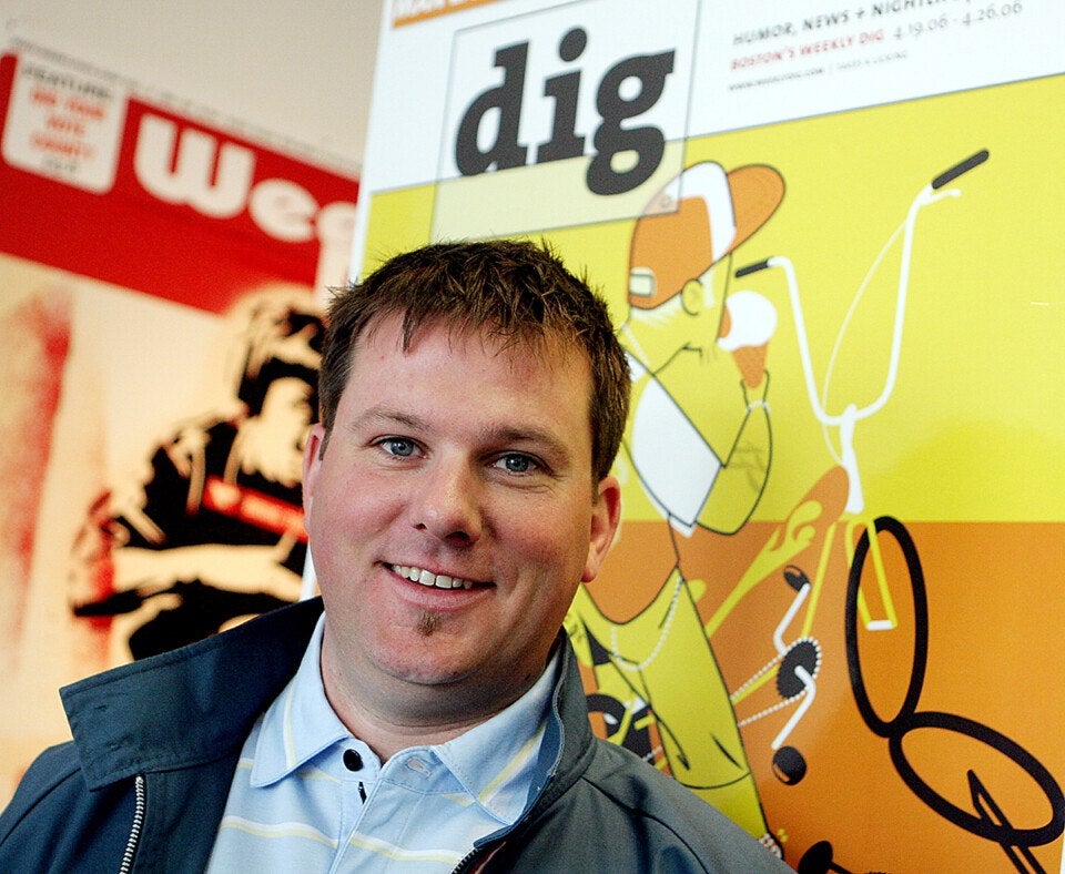 A man with short brown hair and a small patch of facial hair on his chin is smiling. He is wearing a light blue collared shirt with a dark jacket. Behind him is a colorful poster with the word "dig" in bold black letters at the top and an illustration of a person riding a bicycle on a yellow and orange background. Another partially visible poster with red and white colors is in the background.