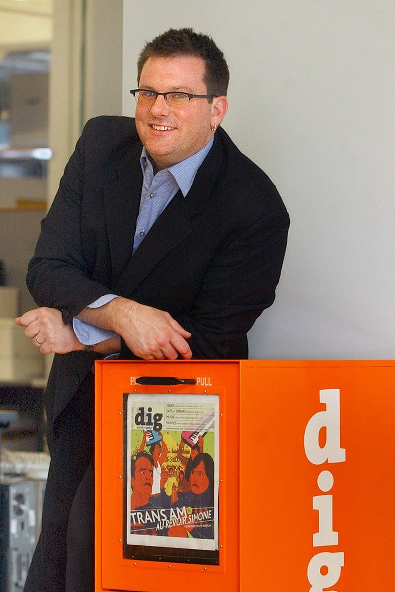 Man wearing glasses, a black blazer, and a light blue shirt leaning on an orange newspaper vending box with the word "dig" written on it and a colorful poster displayed on the front.