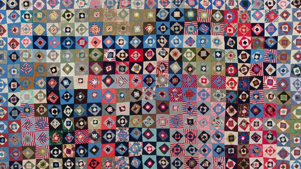 Patchwork quilt composed of numerous small square blocks, each featuring a central diamond shape surrounded by a contrasting border. The blocks display a variety of colors and patterns, including stripes, checks, florals, and solids in shades of blue, red, green, brown, pink, and beige. The overall design is dense and vibrant, with a handmade, vintage appearance.