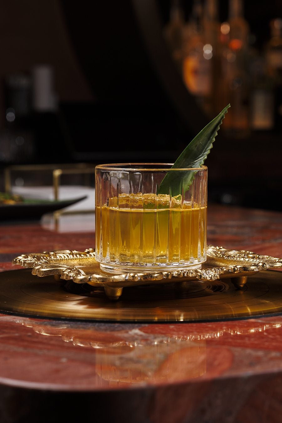 A pale yellow cocktail with one giant ice cube and a palm frond garnish.