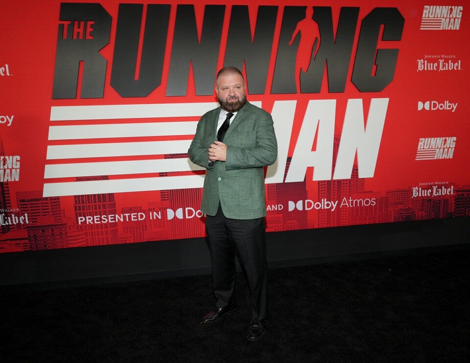 A man with a beard wearing a green blazer, white shirt, black tie, black pants, and black shoes stands on a black carpet in front of a red backdrop with large black and white text that reads "THE RUNNING MAN." The backdrop also features logos for Johnnie Walker Blue Label and Dolby Atmos. The man is looking slightly to his left with his hands clasped in front of him.