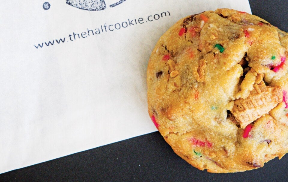 A close-up of a colorful cookie with visible candy pieces and a small waffle cone embedded in it, placed on a white paper with the website "www.thehalfcookie.com" printed on the paper, against a dark background.