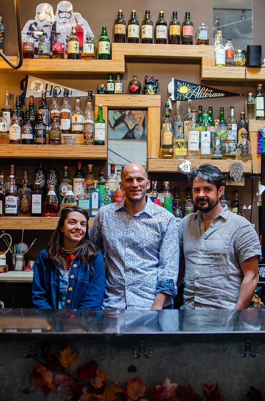 Three people standing behind a bar counter in front of shelves filled with various bottles of liquor. The person on the left is a woman wearing a blue jacket and a red scarf, the person in the middle is a bald man wearing a white shirt with a blue floral pattern, and the person on the right is a man with dark hair and a beard wearing a striped short-sleeve shirt. The shelves behind them are wooden and arranged in a geometric pattern, with a small mirror in the center. There are also decorative items such as a pennant with "Alderaan" written on it and two Stormtrooper cutouts on the top shelf.