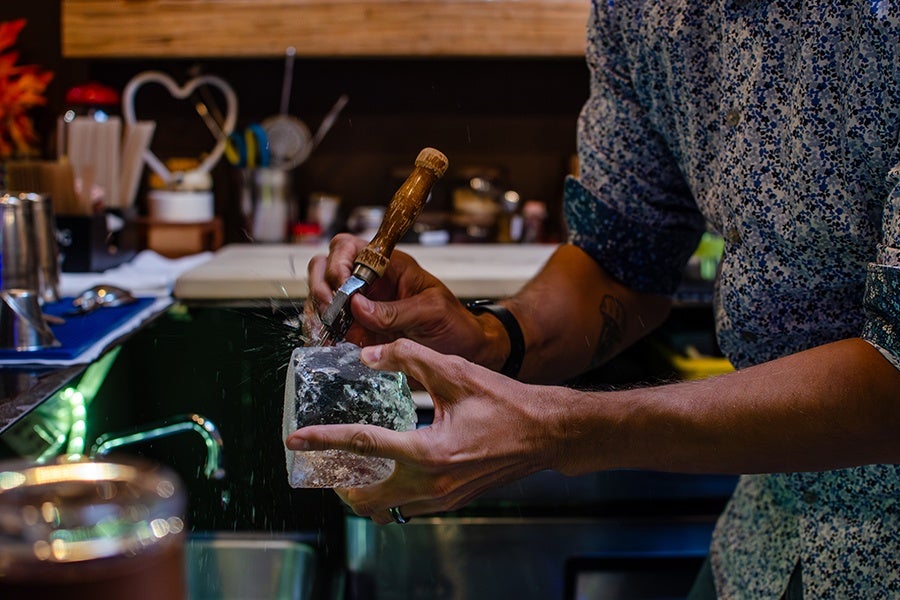 A person wearing a patterned shirt is holding a large block of ice in one hand and using a wooden-handled tool to chip or carve the ice, causing ice shards to scatter. The scene appears to be in a bar or kitchen setting with various utensils and containers in the background.